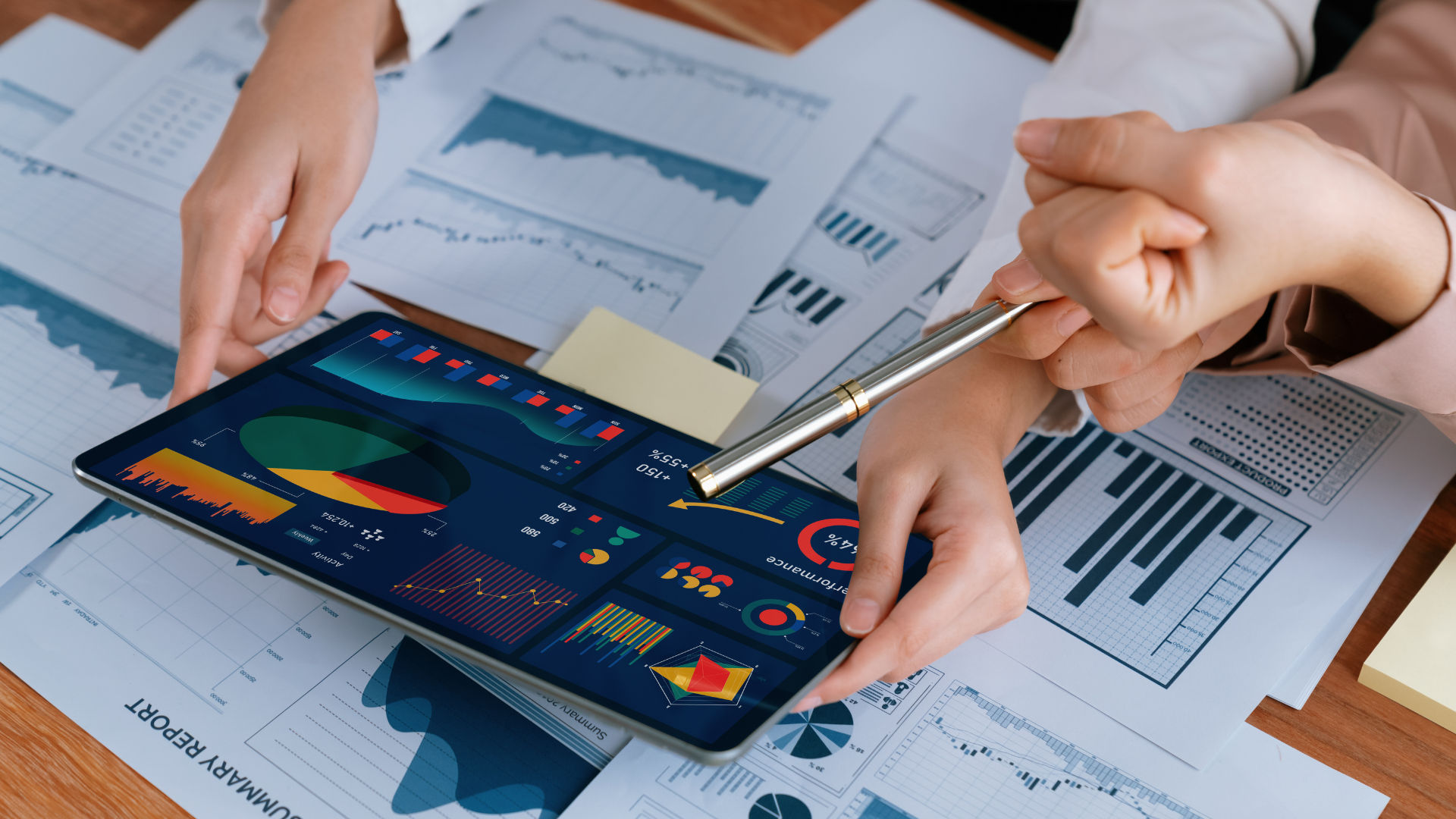 Close-up of hands reviewing financial data on a tablet and printed reports during a financial review meeting, highlighting charts, graphs, and performance metrics