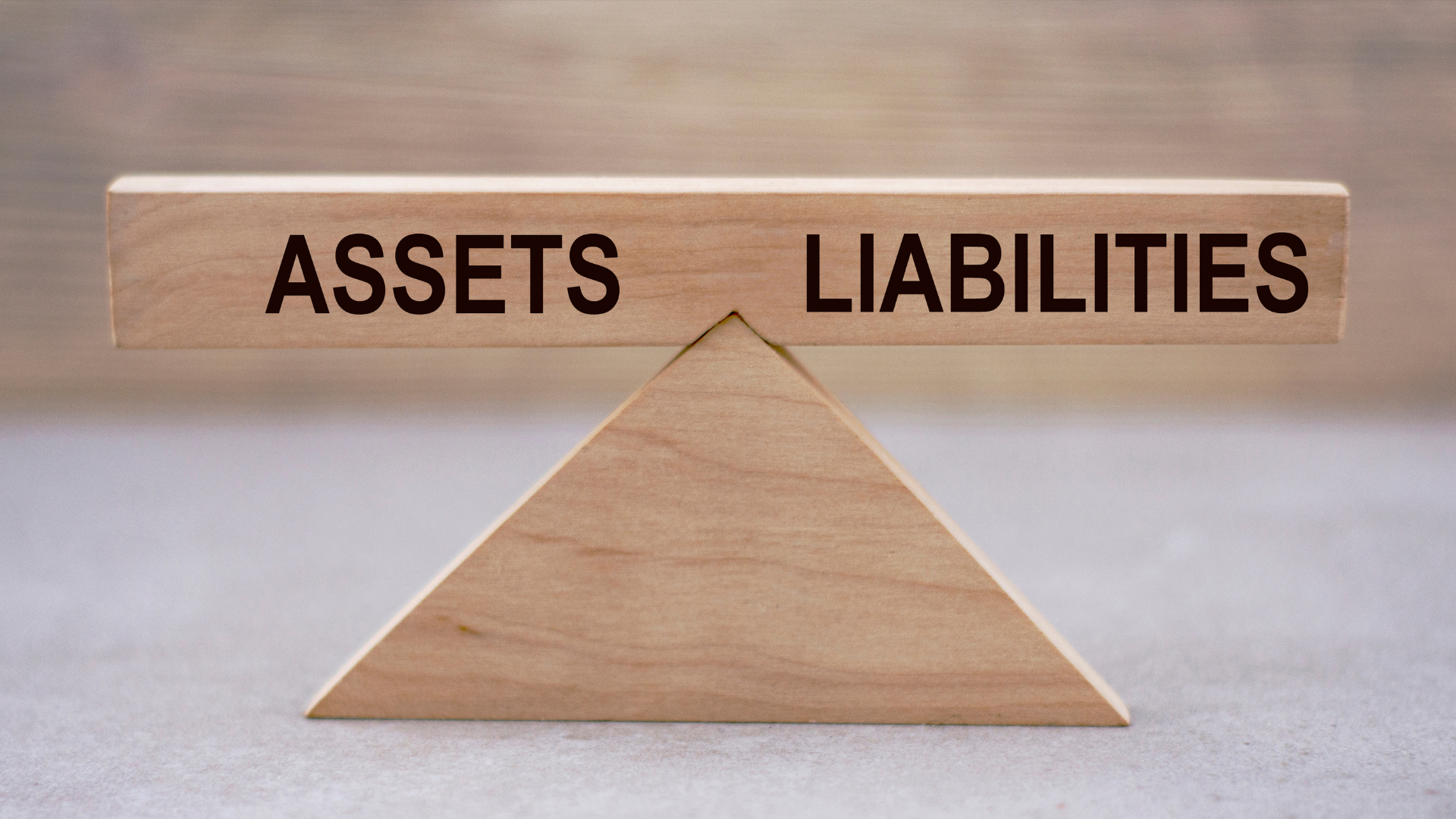 A wooden balance scale with 'Assets' on one side and 'Liabilities' on the other, symbolizing the key components of a Monthly Balance Sheet that provides insights into a business’s financial health and stability.