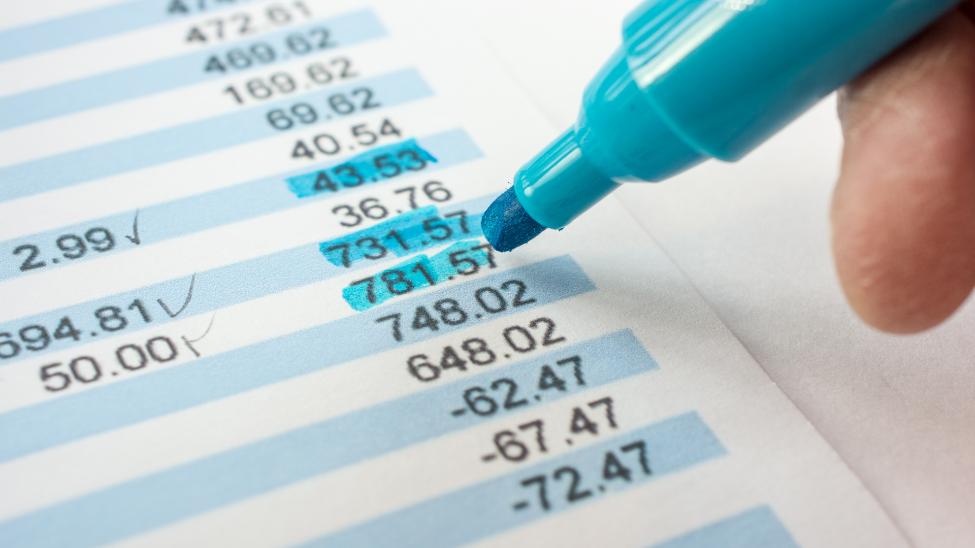 A detailed view of a printed bank statement with alternating light blue and white rows. Specific transaction amounts are highlighted in blue with a marker, and check marks are used to confirm reconciled amounts. A hand holding the blue highlighter is visible, symbolizing the thorough review process involved in Monthly Bank Statement Reconciliations for ensuring financial accuracy.