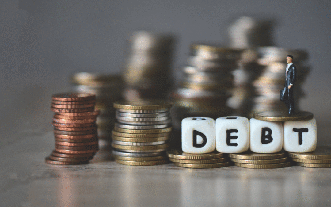 Stacks of coins with the word DEBT spelled out in blocks and a miniature businessperson standing on top, symbolizing the emotional and financial weight of debt for entrepreneurs.